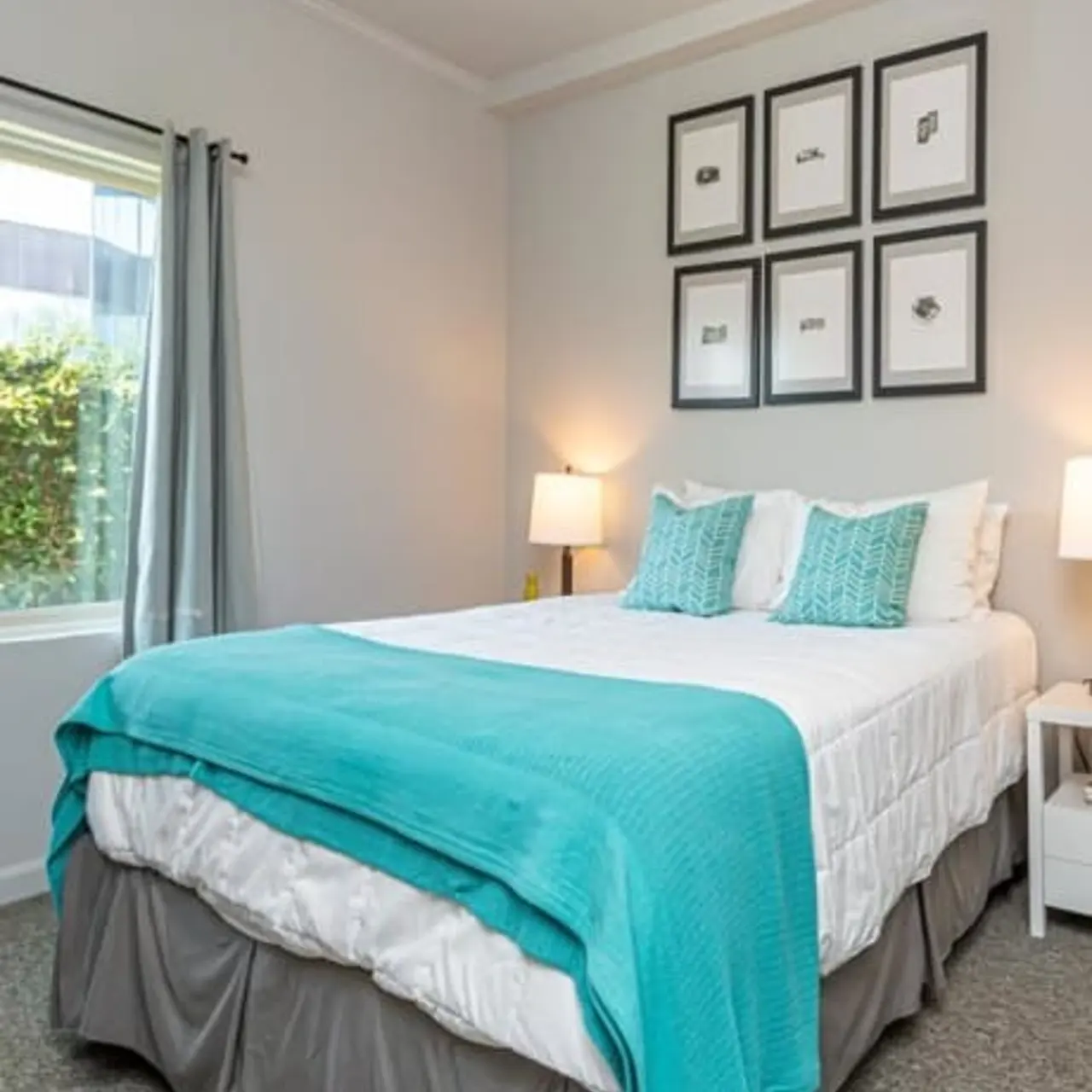 A cozy bedroom featuring a double bed with a turquoise blanket, adorned with two matching decorative pillows. White bedside tables hold lamps, and the walls are decorated with framed art. A window with curtains lets in natural light.