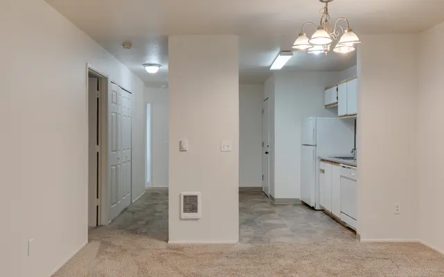 A spacious beige apartment interior showing a living area with carpet, a kitchen visible on the right, and a hallway leading to other rooms on the left.