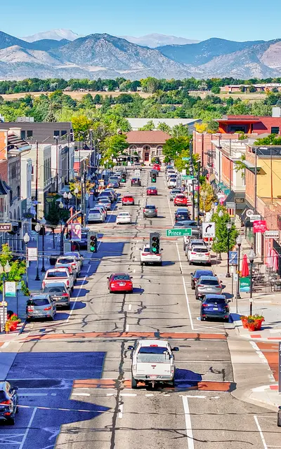 A vibrant downtown street lined with shops and restaurants, featuring parked cars and some pedestrians. In the background, there are mountains and blue skies.
