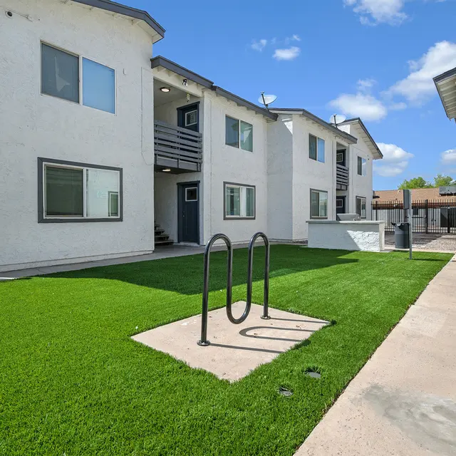 Exterior view of an apartment complex featuring two buildings, a manicured lawn, and a small patio area with a double-sided bicycle rack.