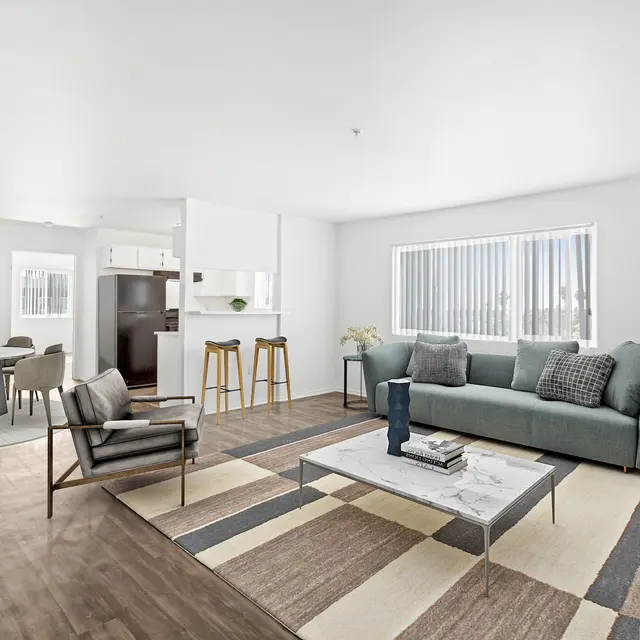 A spacious and modern living room featuring a large sectional sofa, a coffee table, and a patterned area rug. A dining area with gray chairs and a kitchen in the background. Large windows allow natural light to fill the room, complemented by indoor plants.