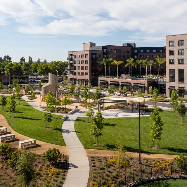 A modern urban park featuring a children's playground, walking paths, and green lawns bordered by new buildings.