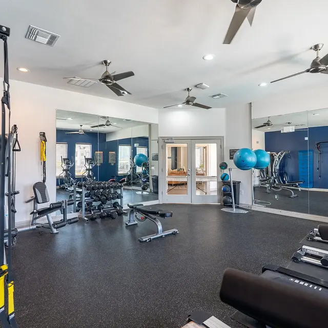 A spacious gym with various exercise equipment including weight machines, benches, and exercise balls, surrounded by mirrors and ceiling fans.