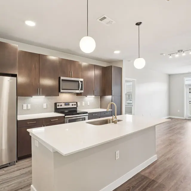A modern kitchen featuring dark wood cabinets, stainless steel appliances, and a large island. The space is bright with pendant lights and a view of a living area with access to a balcony.