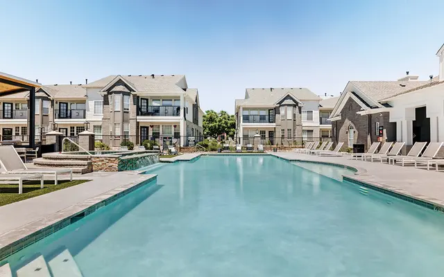 A spacious outdoor pool area surrounded by residential buildings, featuring lounge chairs and a clear blue sky overhead.