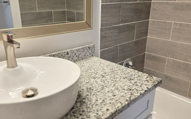Modern Bathroom Design A contemporary bathroom featuring a round sink on a granite countertop, with a bath tub and a mirror in the background. The walls are adorned with gray tiles.
