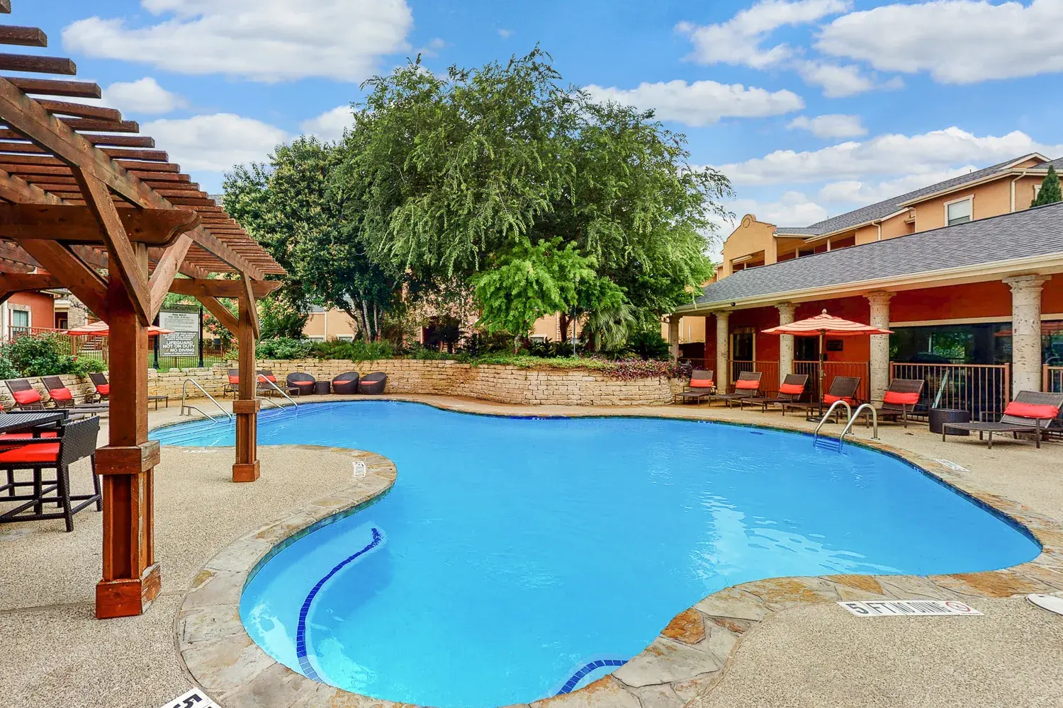A serene swimming pool area featuring a blue pool surrounded by a stone edge, a covered patio, and lush greenery in the background.