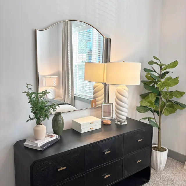 A modern bedroom dresser with a large mirror above it. The dresser features multiple drawers and decorative items like books and a small plant. A stylish lamp with a spiral design is illuminated next to the dresser, accompanied by a potted plant in the corner.
