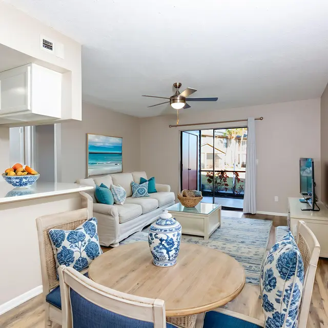 A bright and modern living area featuring a comfortable white sofa, a round wooden table with blue accents, and a sliding glass door leading to a balcony. The room has light walls, wooden flooring, and decorative elements including a fruit bowl and a blue vase.