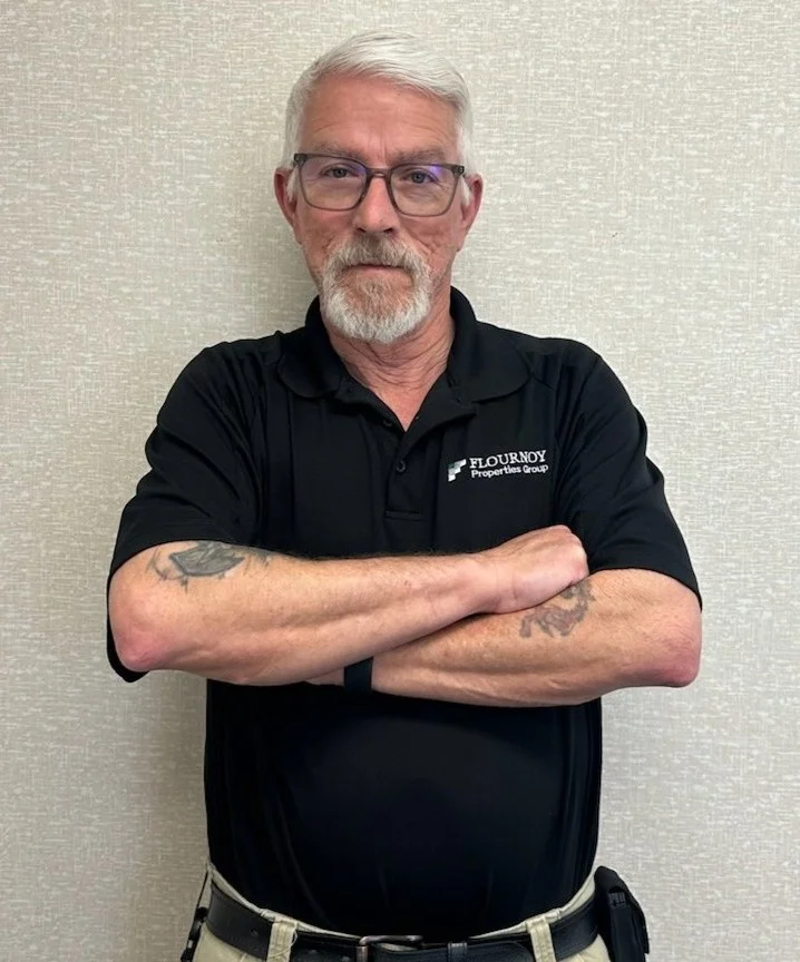 Confident Portrait of a Man A man with silver hair and glasses stands with his arms crossed against a neutral beige wall. He wears a black polo shirt embroidered with the name 'Flournoy'.