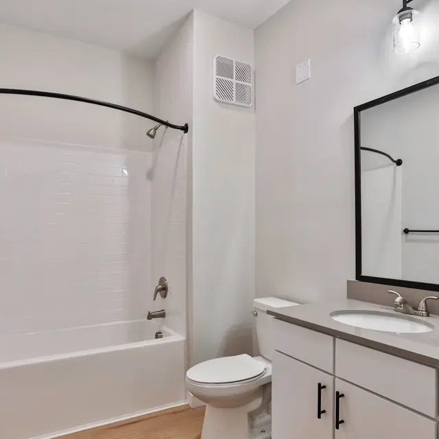 A modern bathroom featuring a white bathtub with a black curtain rod, a large mirror above a white vanity with silver fixtures, and light wood flooring.