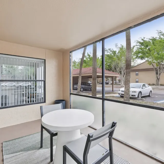 An outdoor screened patio with a round table and chairs, surrounded by trees and a parking area.