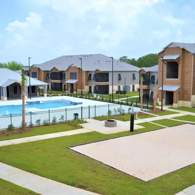 A modern apartment complex featuring two buildings surrounding a swimming pool, with green lawns and a volleyball court visible. The sky is partly cloudy, creating a pleasant outdoor atmosphere.