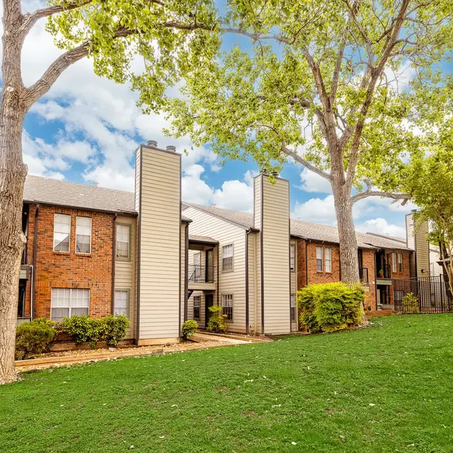 View of a brick apartment complex surrounded by green grass and trees under a partly cloudy sky.