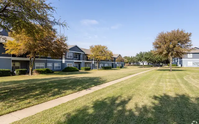 A landscaped apartment complex with multiple gray buildings, featuring a well-maintained lawn and walking path under a clear blue sky.