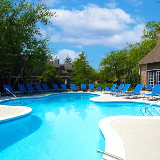A serene swimming pool area surrounded by lounge chairs and lush greenery under a blue sky with clouds.