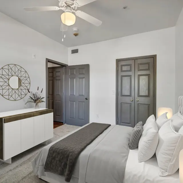A modern bedroom featuring a bed with white bedding, a white nightstand, and a potted plant. There are two gray doors and wall art depicting leaves.