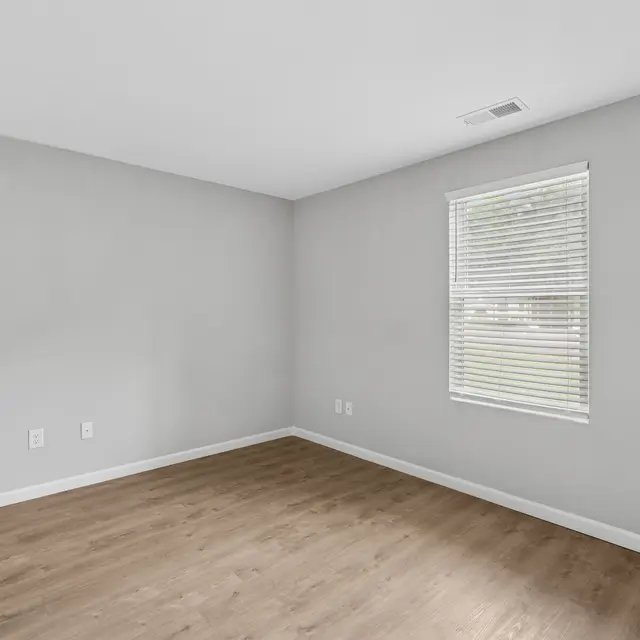 An empty room with light grey walls and wood-like flooring. A window with blinds allows natural light to enter.