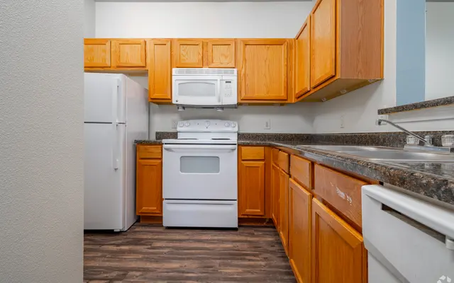 A modern kitchen featuring wooden cabinets, a white refrigerator, and a white stove with microwave above it. Countertops have a dark granite finish, and there's a double sink with a chrome faucet.
