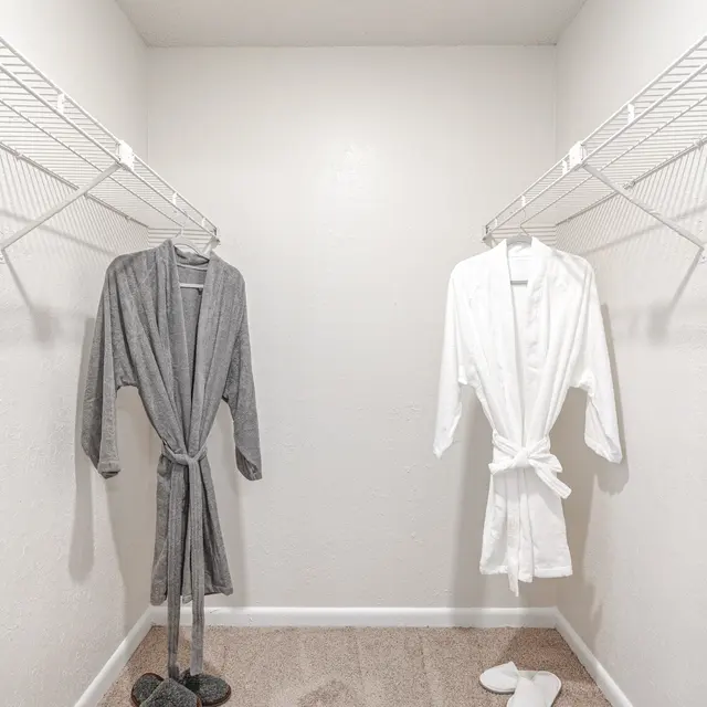 An interior view of a closet with two hanging robes—one gray and one white—on wire shelves. The floor is carpeted and there are slippers in front of the robes.