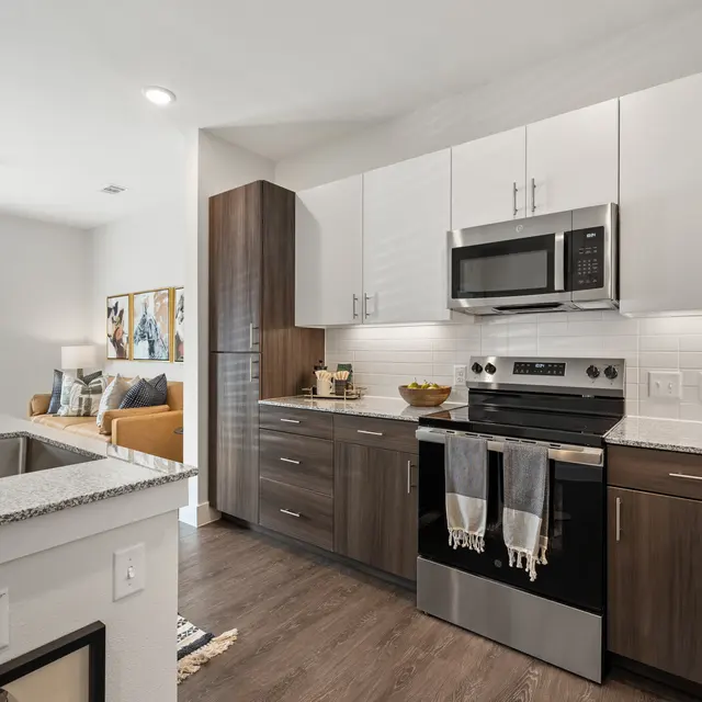 A modern kitchen with dark cabinets and stainless steel appliances, featuring a view into a cozy living area with a sofa and decorative artwork.