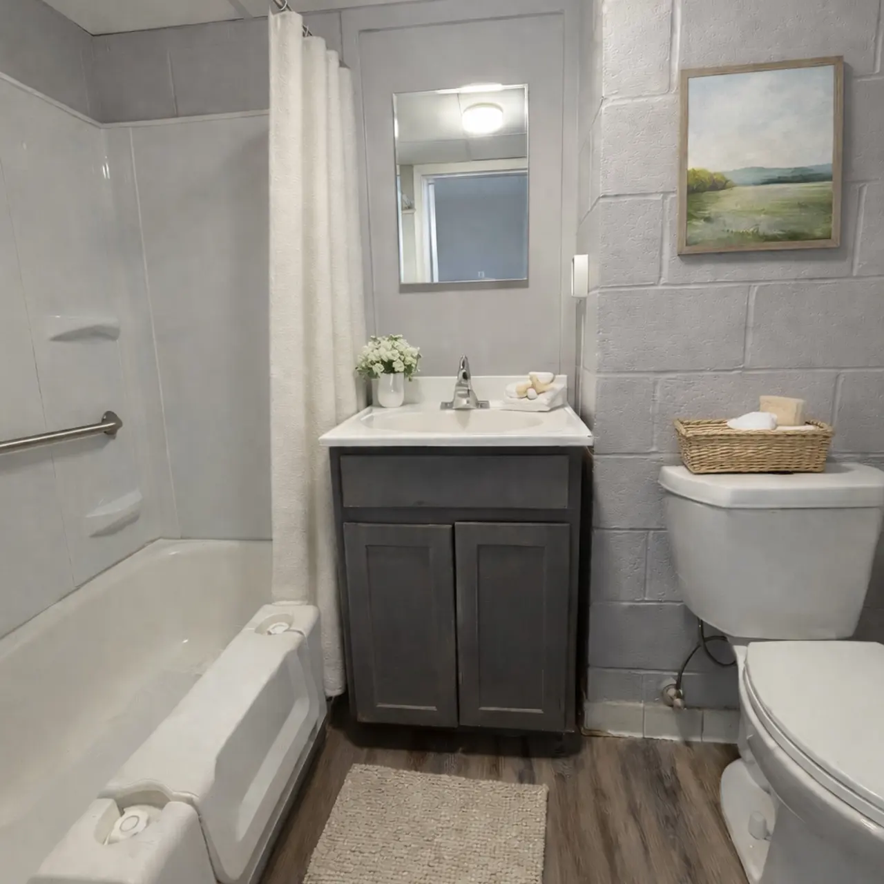 A modern bathroom featuring a bathtub with a shower curtain, a small sink vanity, a mirror, and a toilet. The space has gray walls, wooden flooring, and decorative items such as a flower vase and a framed painting.