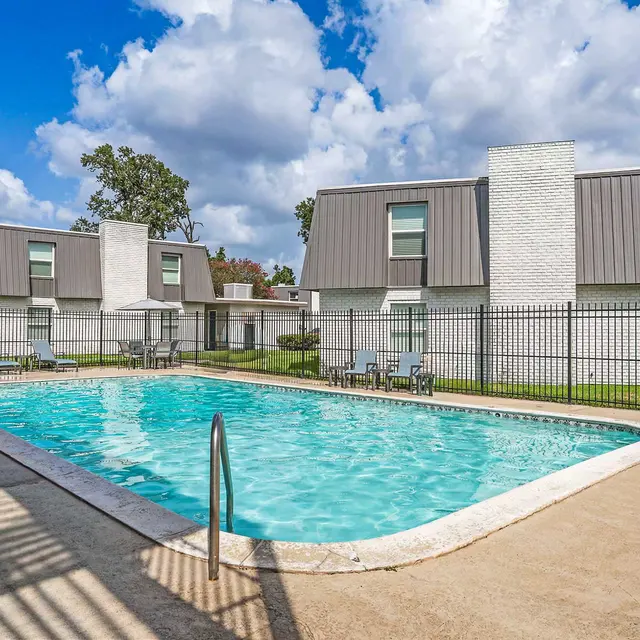 A clear blue swimming pool in an apartment complex, surrounded by fenced greenery and residential buildings with sloped roofs under a partly cloudy sky.