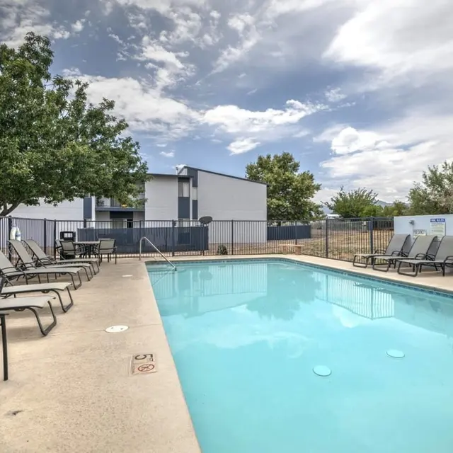 A clear swimming pool surrounded by lounge chairs and a fenced area, with trees and a building in the background under a cloudy sky.