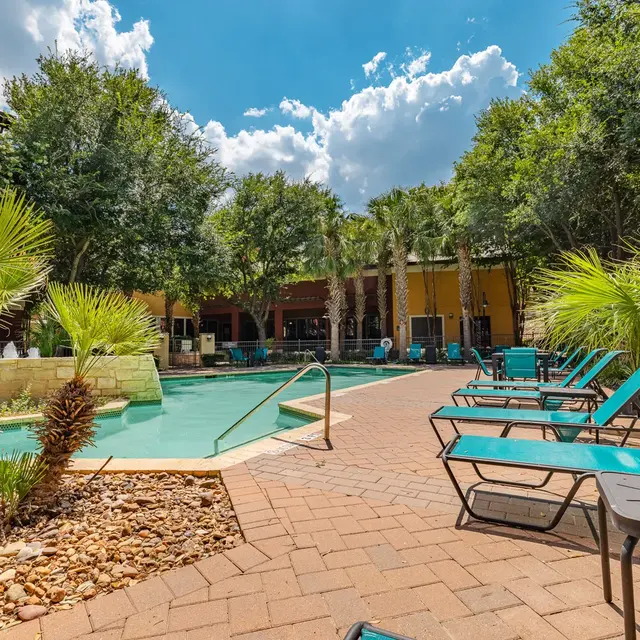 A sunny pool area with lounge chairs and palm trees, surrounded by greenery and a building in the background.