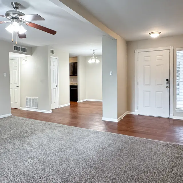 Spacious living room featuring light-colored walls, a ceiling fan, and carpet flooring, leading to a door and two closets.