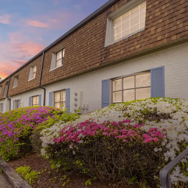 Exterior view of a cozy apartment building with vibrant flowering shrubs in the foreground and a lamppost on the path, under a colorful sunset sky.