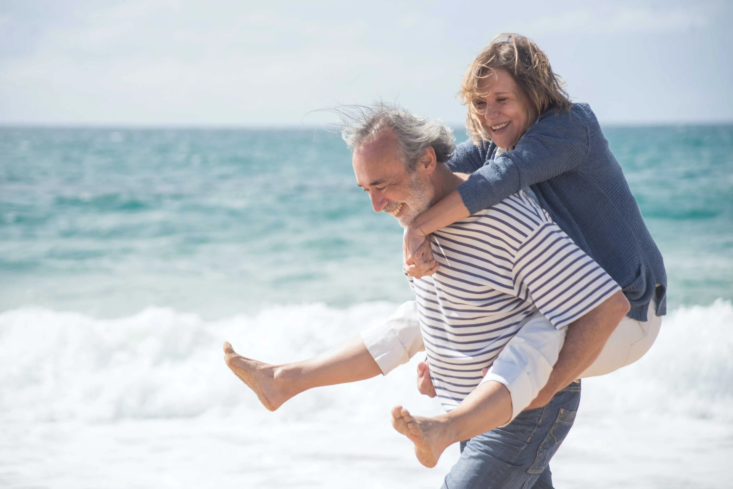 A joyful couple enjoying time at the beach, with the man carrying the woman on his back. They are smiling and playing in the surf with waves in the background.