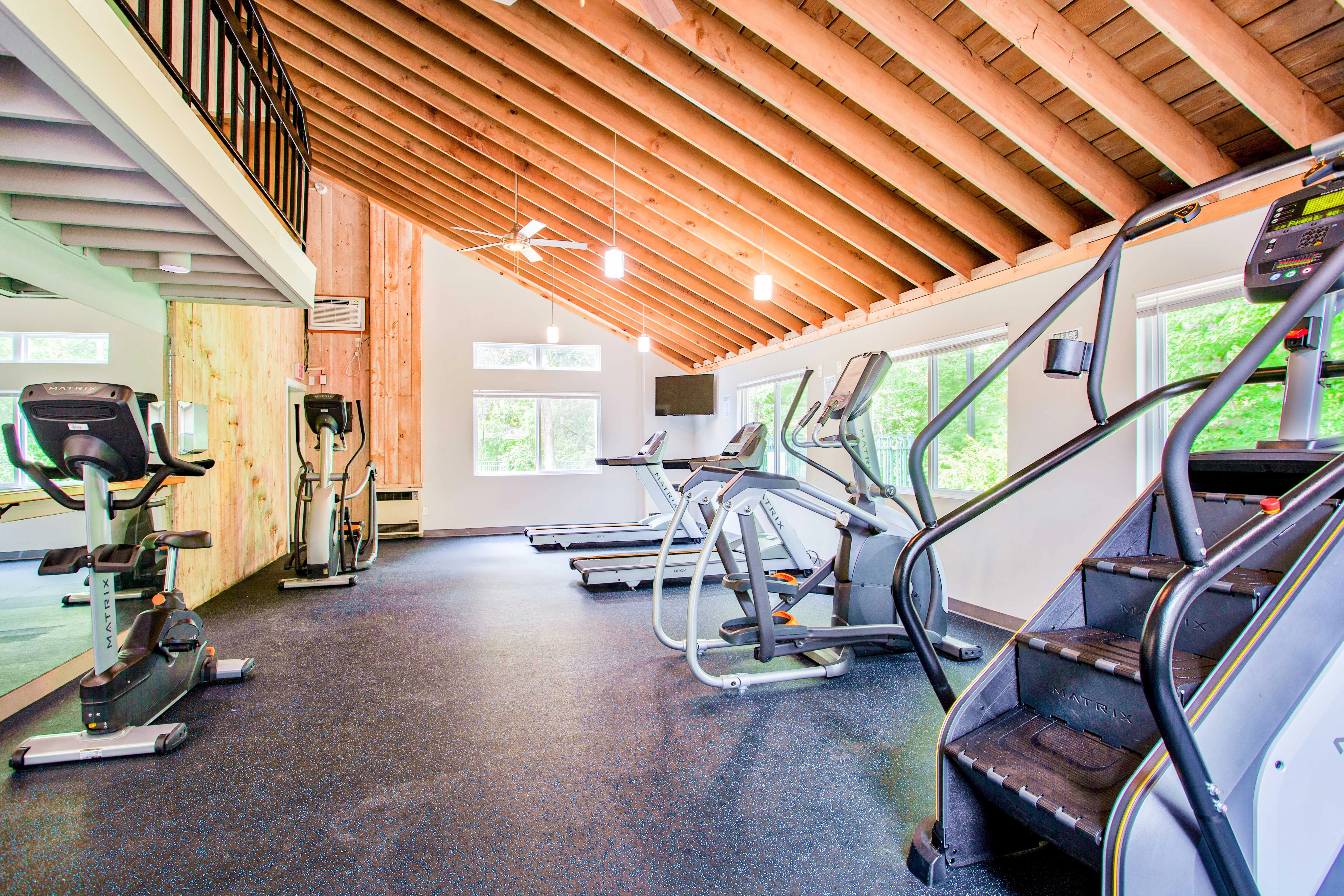 Interior view of a modern gym with wooden ceilings and large windows, featuring various exercise equipment including treadmills, stationary bikes, and an elliptical machine.