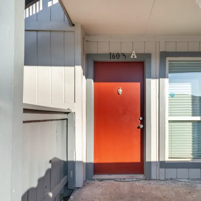 Close-up view of a red apartment door with a door number and a small window beside it, surrounded by a gray exterior wall and balcony.