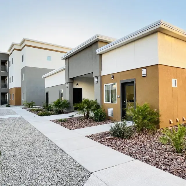 A modern apartment complex featuring two-story buildings with varying colors, landscaped pathways, and decorative shrubs.