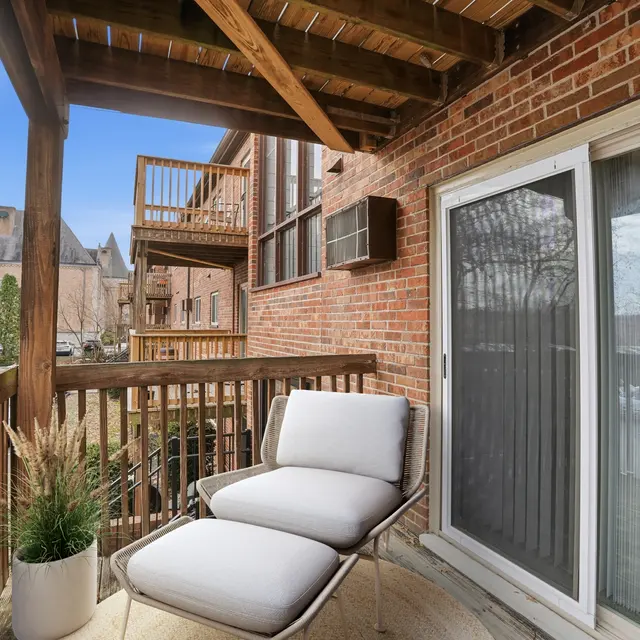 A cozy balcony with a modern lounge chair and potted plant, featuring wooden railings and brick walls, with a view of neighboring buildings.