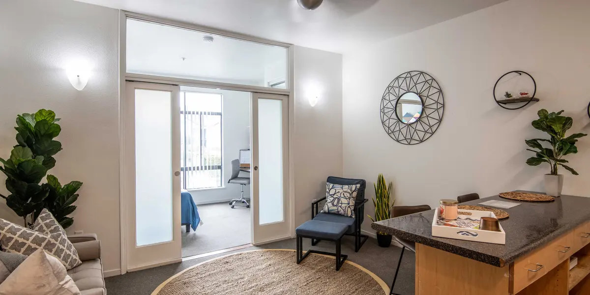 Modern living room with a sofa, round rug, plants, and doorway leading to another room.