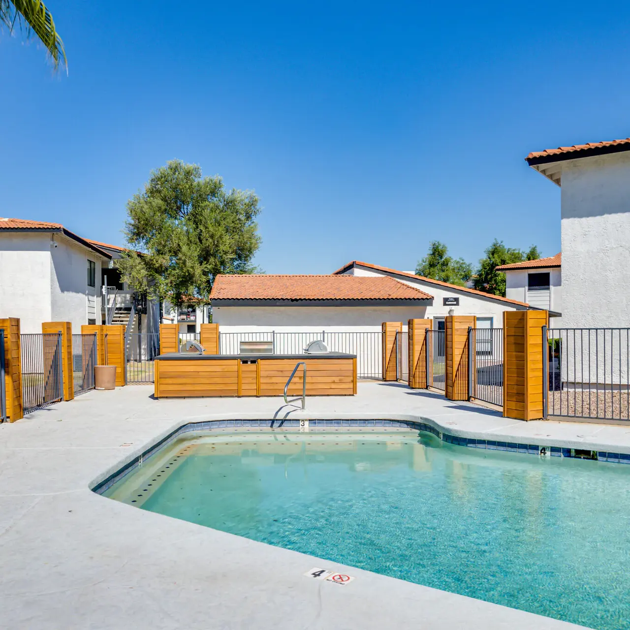 A clean swimming pool area surrounded by buildings in a residential complex, with blue skies and palm trees in the background.
