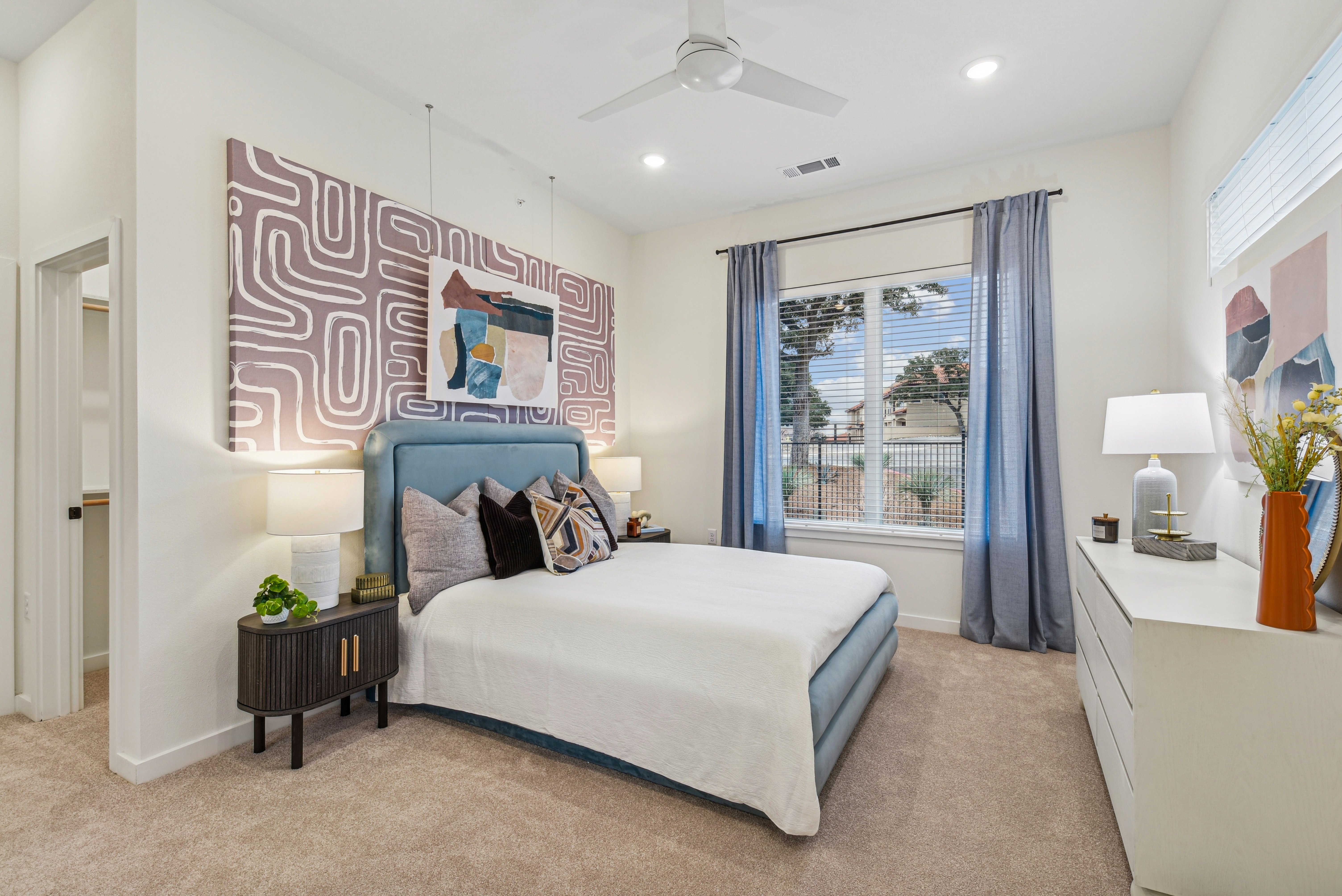 A contemporary bedroom featuring a blue upholstered bed, decorative pillows, and art on the wall. Natural light filters through sheer curtains over a window, accompanied by a modern dresser and nightstand with lamps and decor elements.