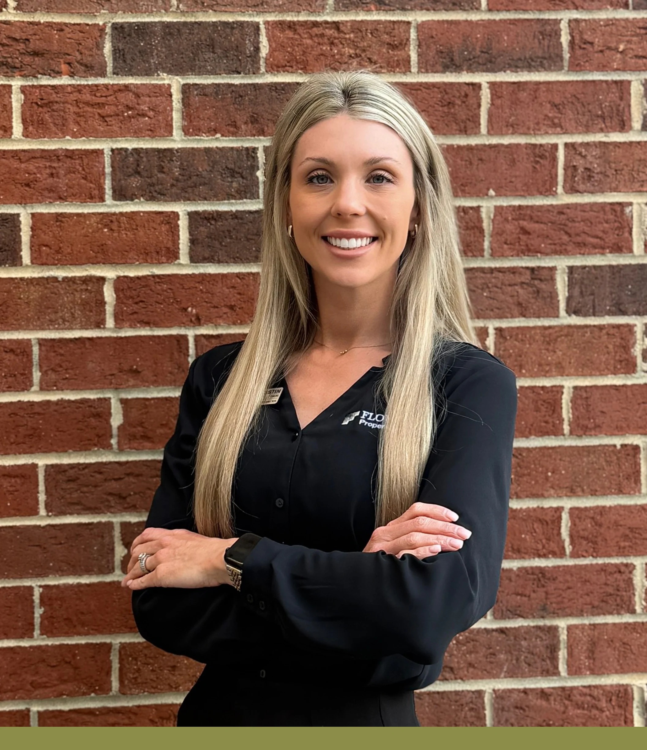 Professional Portrait in Front of Brick Wall A woman standing in front of a brick wall, wearing a black blouse with arms crossed and smiling confidently.