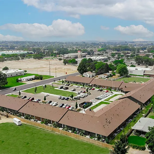 Aerial View of Apartment Complex Aerial view of a spacious apartment complex surrounded by greenery and parking areas, with a backdrop of distant buildings and blue skies.