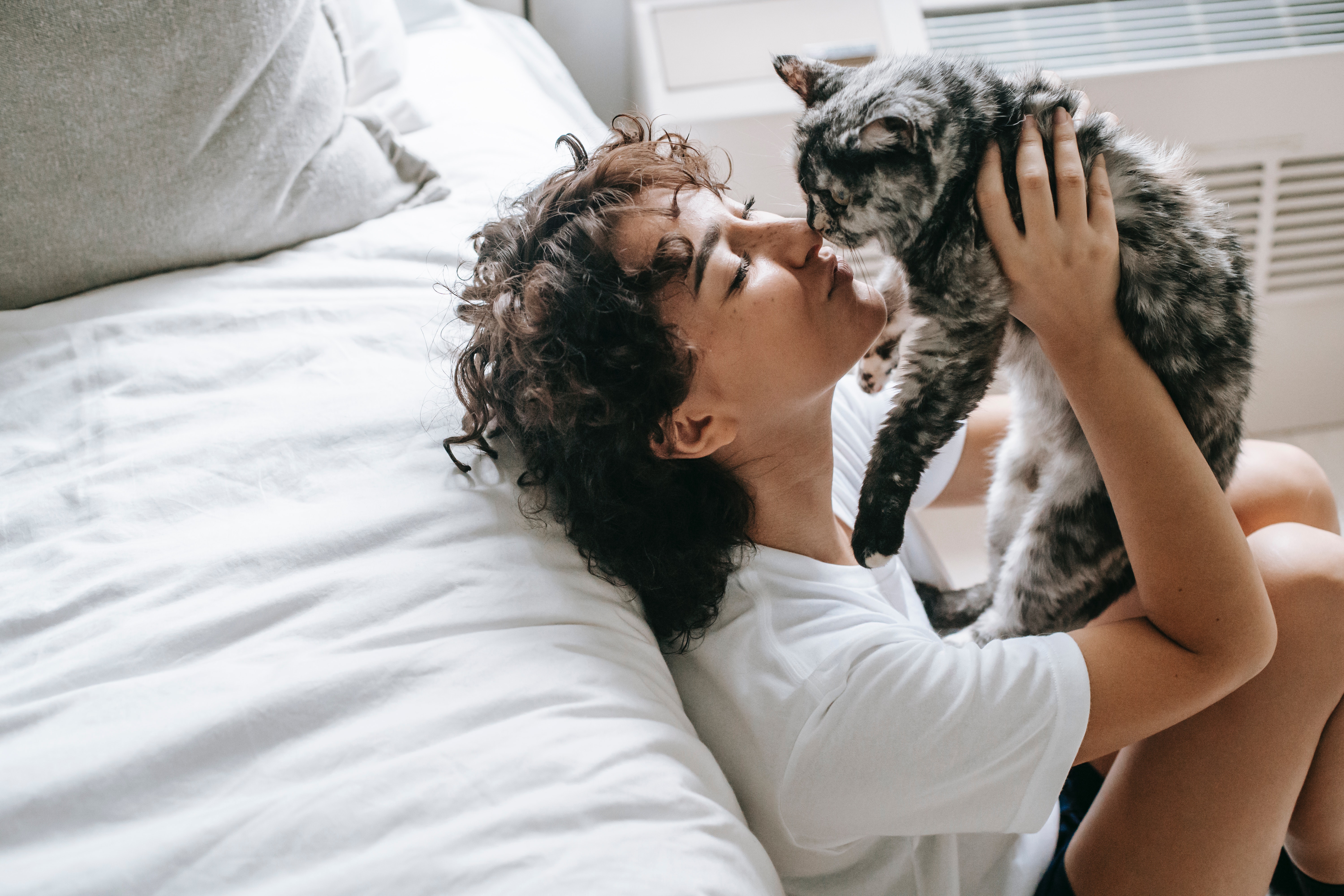 A Woman Kissing Her Cat A woman with curly hair sitting on a bed, affectionately kissing her gray cat as she holds it close.
