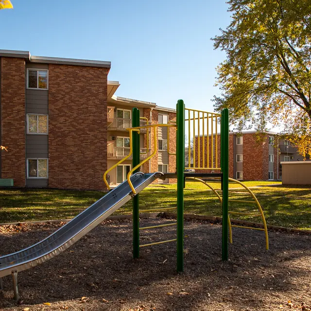 A playground featuring a slide and climbing structure set against a backdrop of brick apartment buildings under a clear blue sky.