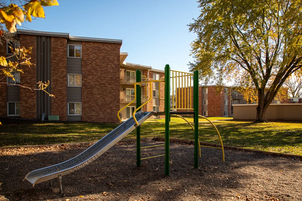 A playground featuring a slide and climbing structure set against a backdrop of brick apartment buildings under a clear blue sky.