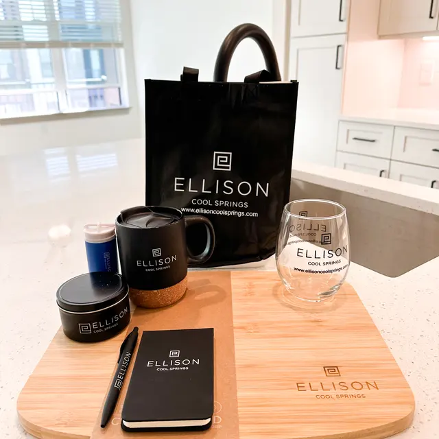 A collection of branded items on a wooden serving board, including a black mug, glass, notebook, pen, scented candle, and a black tote bag with 'ELLISON COOL SPRINGS' logo.