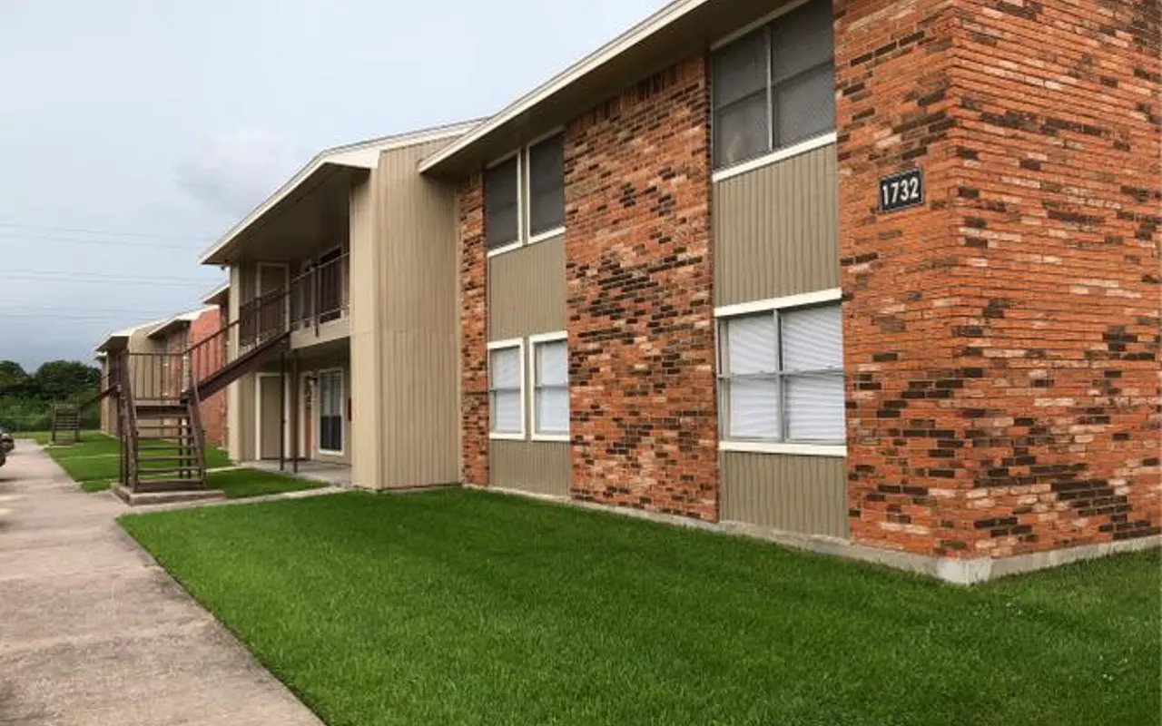 Exterior view of a two-story apartment complex with a mix of brick and siding, featuring green grass and a concrete pathway.