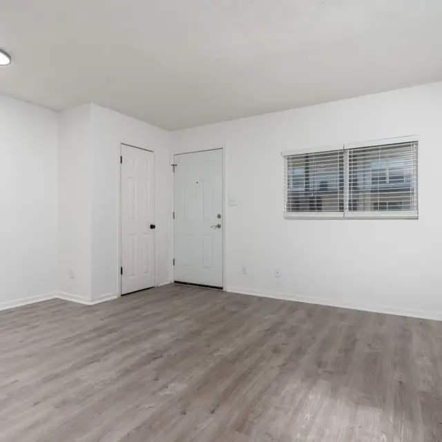 An empty room with light-colored walls and wooden flooring; features a door on the left and a window on the right.