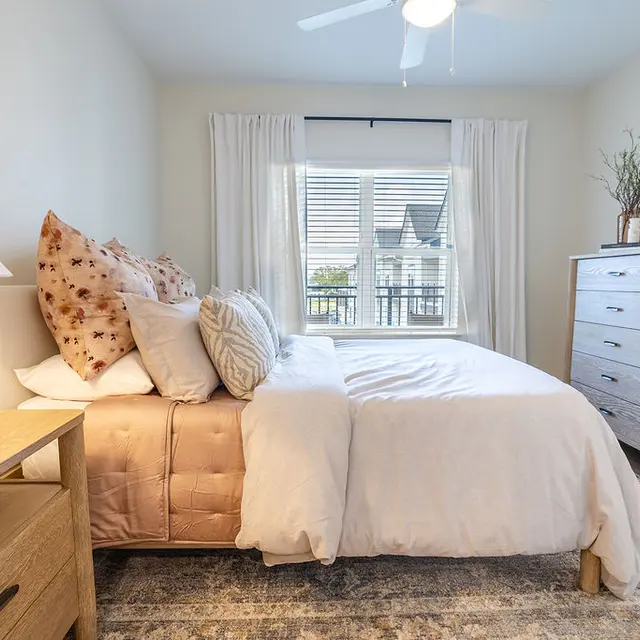 A cozy bedroom featuring a bed with textured pillows, light-colored bedding, and a bedside lamp. A dresser stands against the wall, and a window with sheer curtains allows natural light to enter the room.