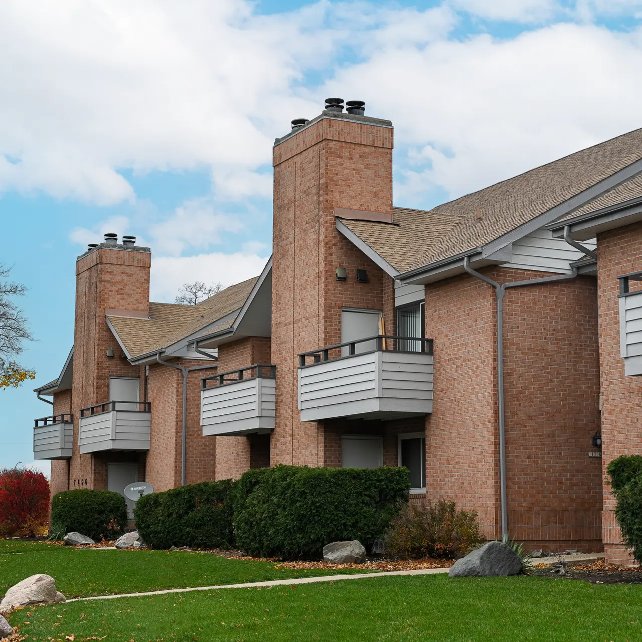 A photo of a brick apartment complex with two-story buildings featuring balconies. The area is landscaped with bushes and stones, under a cloudy sky.