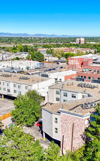 Aerial view of a suburban city with various buildings, parking lots, and greenery. The landscape includes low-rise buildings with rooftops visible, separated by streets and patches of trees. In the background, mountains can be seen under a clear blue sky.
